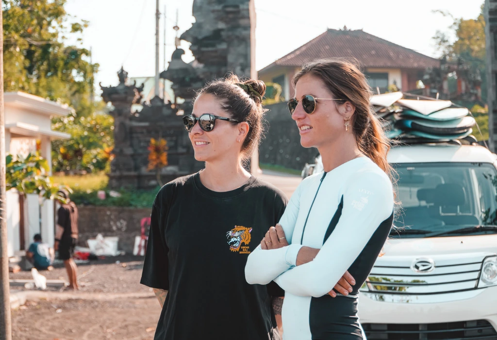 Two girls looking towards the beach with sunglasses on in front of a car with surfboards on the roof